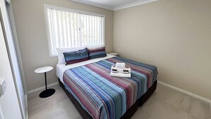 Un dormitorio con cama, mesita de noche, ventana con persianas y una pequeña mesa redonda.