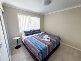 A bedroom with a bed, a bedside table, a window with blinds, and a small round table.