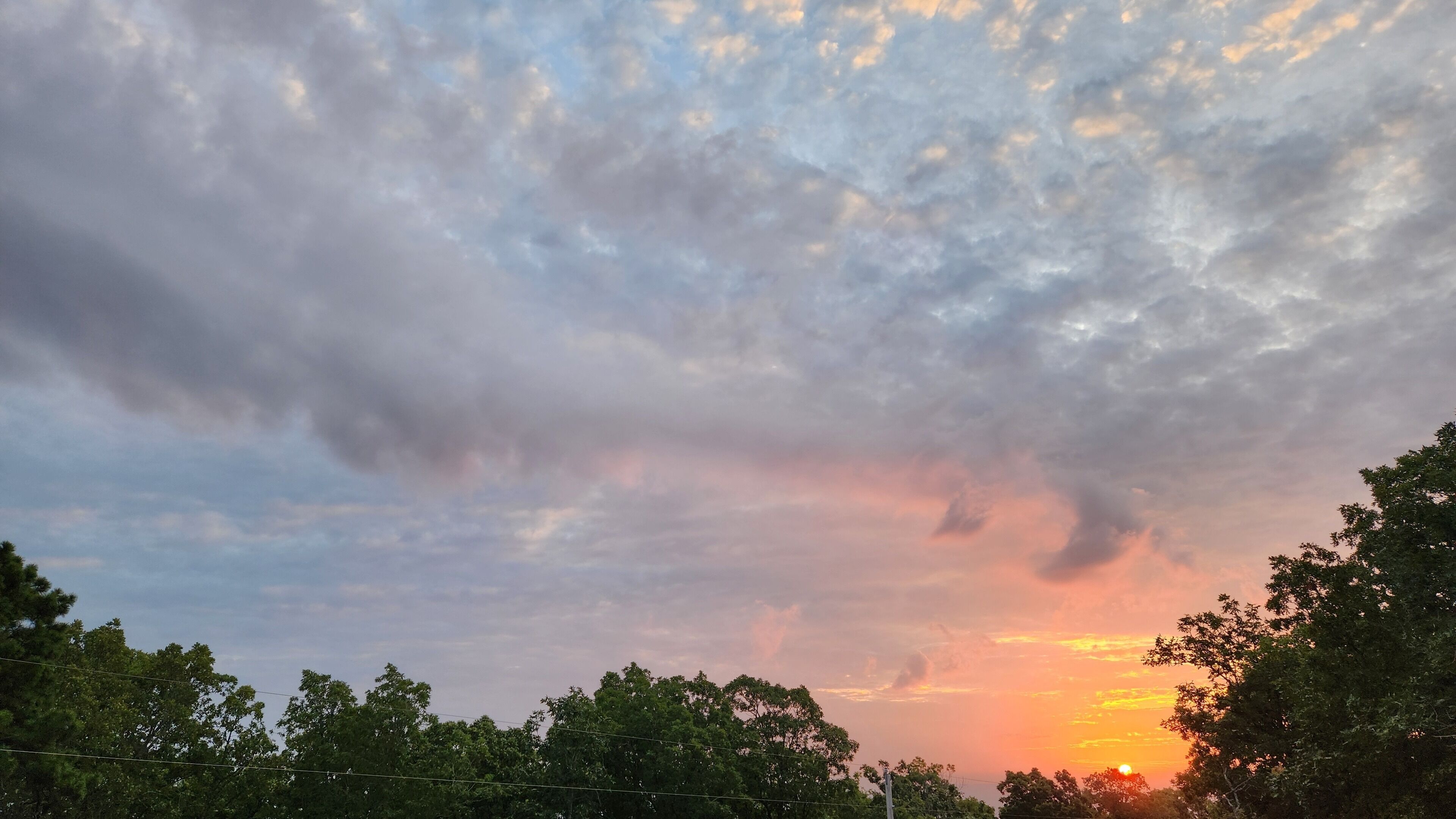 Een zonsondergang met wolken en bomen op de voorgrond.