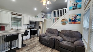 A modern kitchen with white cabinets, a stainless steel refrigerator, and a microwave. There is a wooden floor and a gray sofa in the living area.