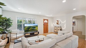 Una sala de estar moderna con un televisor de pantalla plana, sofás blancos, una mesa de centro y un ventanal con persianas.