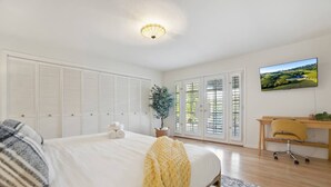 Un dormitorio con cama, escritorio, silla, planta y un cuadro en la pared.