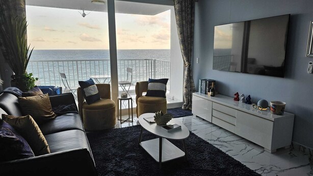 Ein modernes Wohnzimmer mit großem Fenster mit Meerblick, einer Sofagarnitur, einem Couchtisch und einem Fernseher an der Wand.