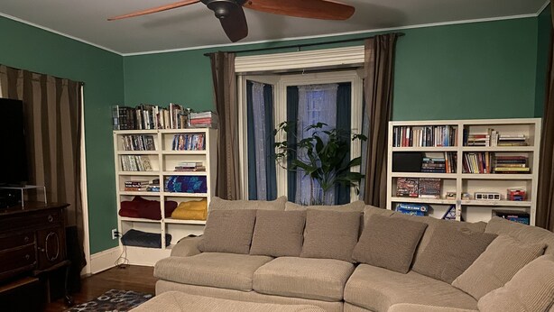 Een woonkamer met een hoekbank, een plafondventilator, een boekenkast en een televisie.