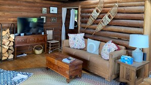 Un acogedor interior de cabaña de madera con sofá, mesa de centro de madera, sillas de mimbre, televisor y chimenea.