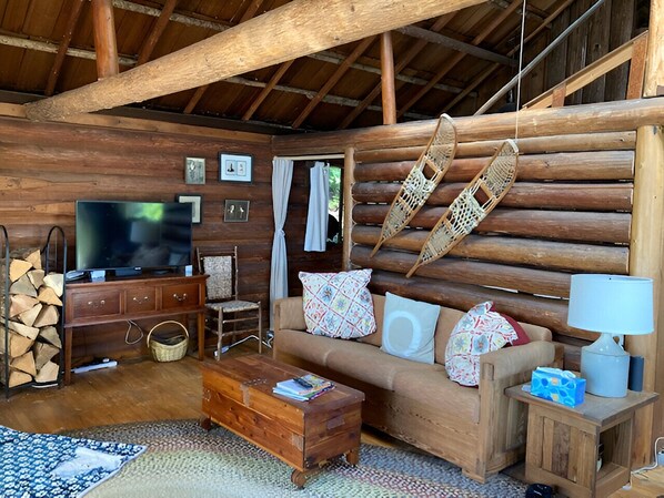 Un acogedor interior de cabaña de madera con sofá, mesa de centro de madera, sillas de mimbre, televisor y chimenea.