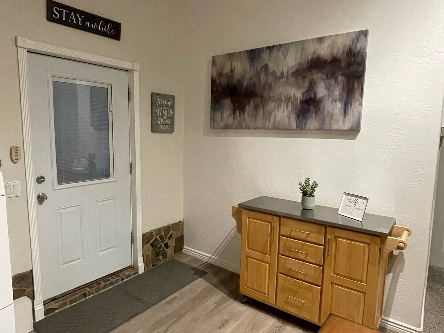 A reception area with a white door, a stone accent wall, a wooden cabinet with drawers, a potted plant, and a sign that says "STAY awhile".