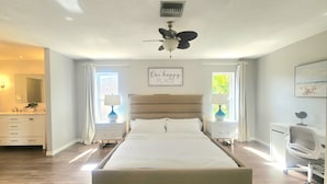 A bedroom with a large bed, a ceiling fan, a window with curtains, a bedside table with a lamp, and a wall-mounted artwork. - BEACH GETAWAY- 9 MIN FROM THE BEACH (Bonita Springs)