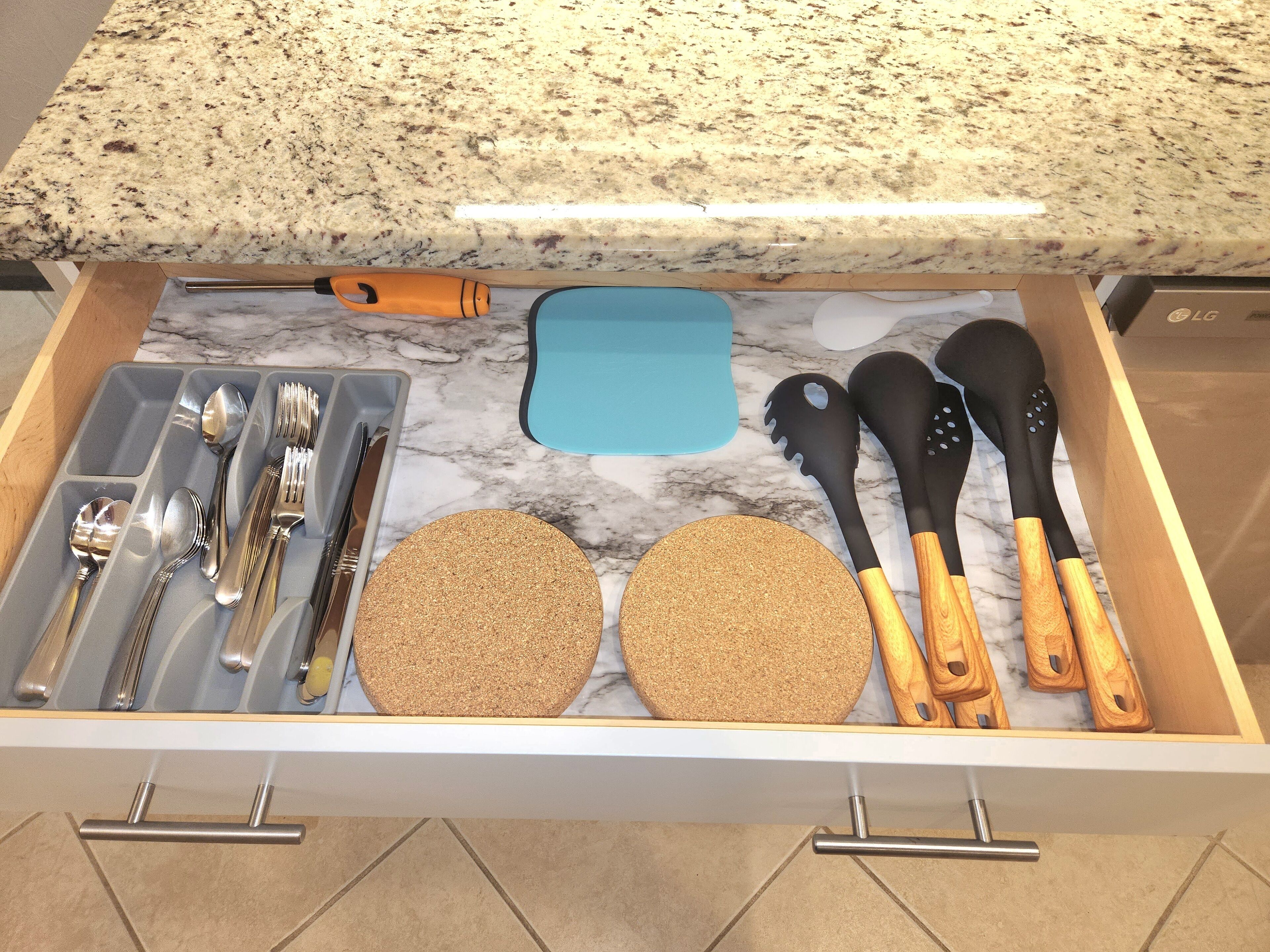 Un cajón de cocina con utensilios, una tabla de cortar y posavasos de corcho.