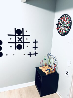 A room with a dartboard, a tic-tac-toe game, and a wooden toy on a black side table.
