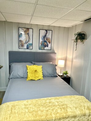 A neatly made bed with a yellow accent pillow, a headboard with a decorative panel, and two framed artworks above the bed.