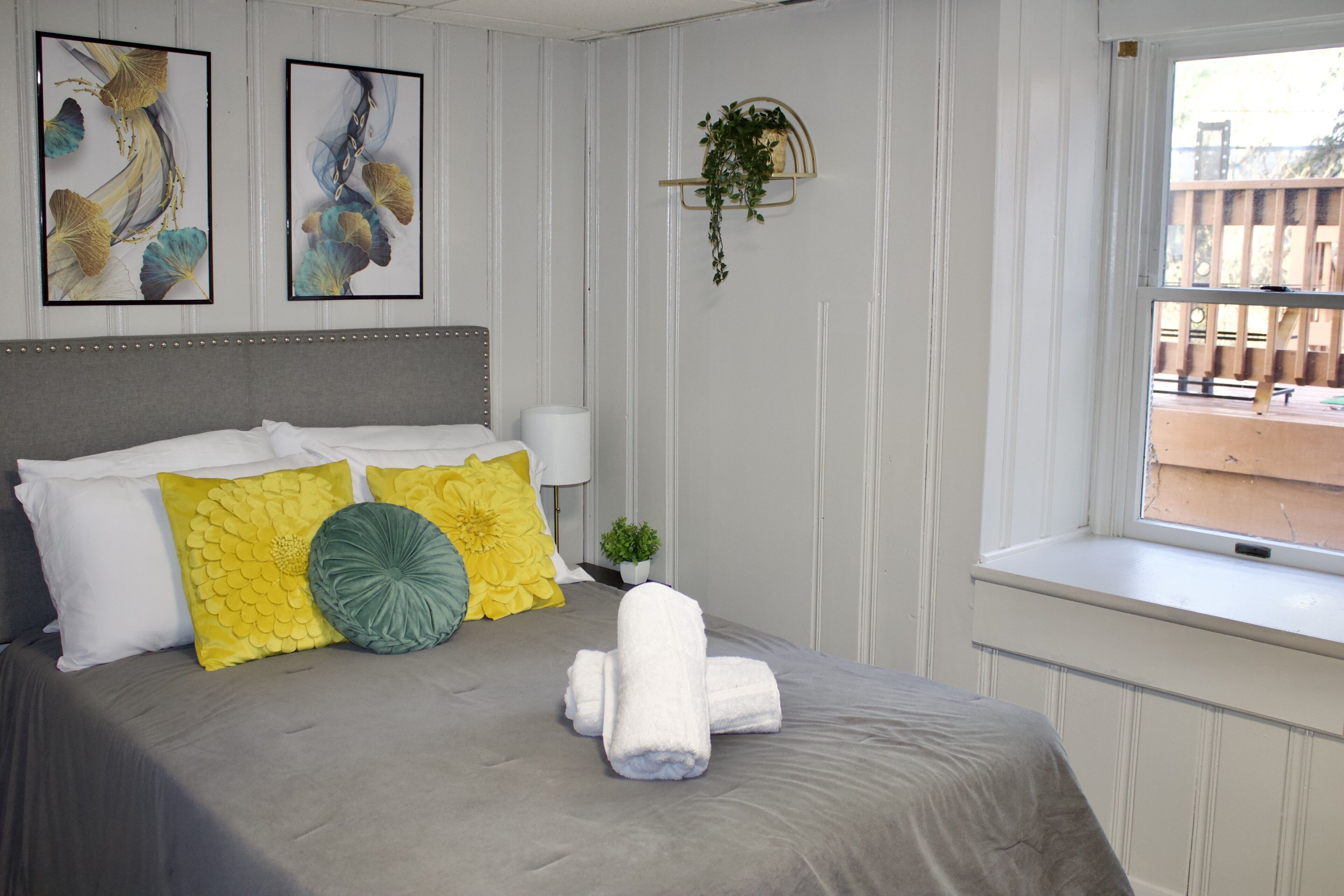 A neatly made bed with a grey bedspread, white pillows, and a yellow and green decorative pillow. There are two framed artworks on the wall above the bed, and a small potted plant hanging on the wall.