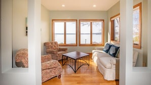 A living room with a wooden floor, two armchairs, a white sofa, and a coffee table. Large windows offer a view of a body of water.