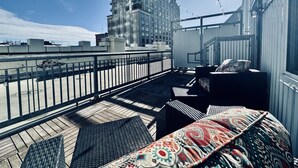 Una terraza en la azotea con vistas a un edificio de gran altura, mobiliario exterior e iluminación con luces colgantes.