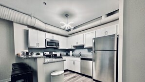 Una cocina moderna con armarios blancos, electrodomésticos de acero inoxidable, horno integrado, microondas y refrigerador. El piso es de madera y hay un bote de basura.