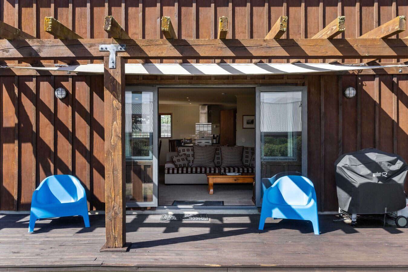 A wooden building with a covered entrance, two blue chairs, a barbecue grill, and a sofa inside.