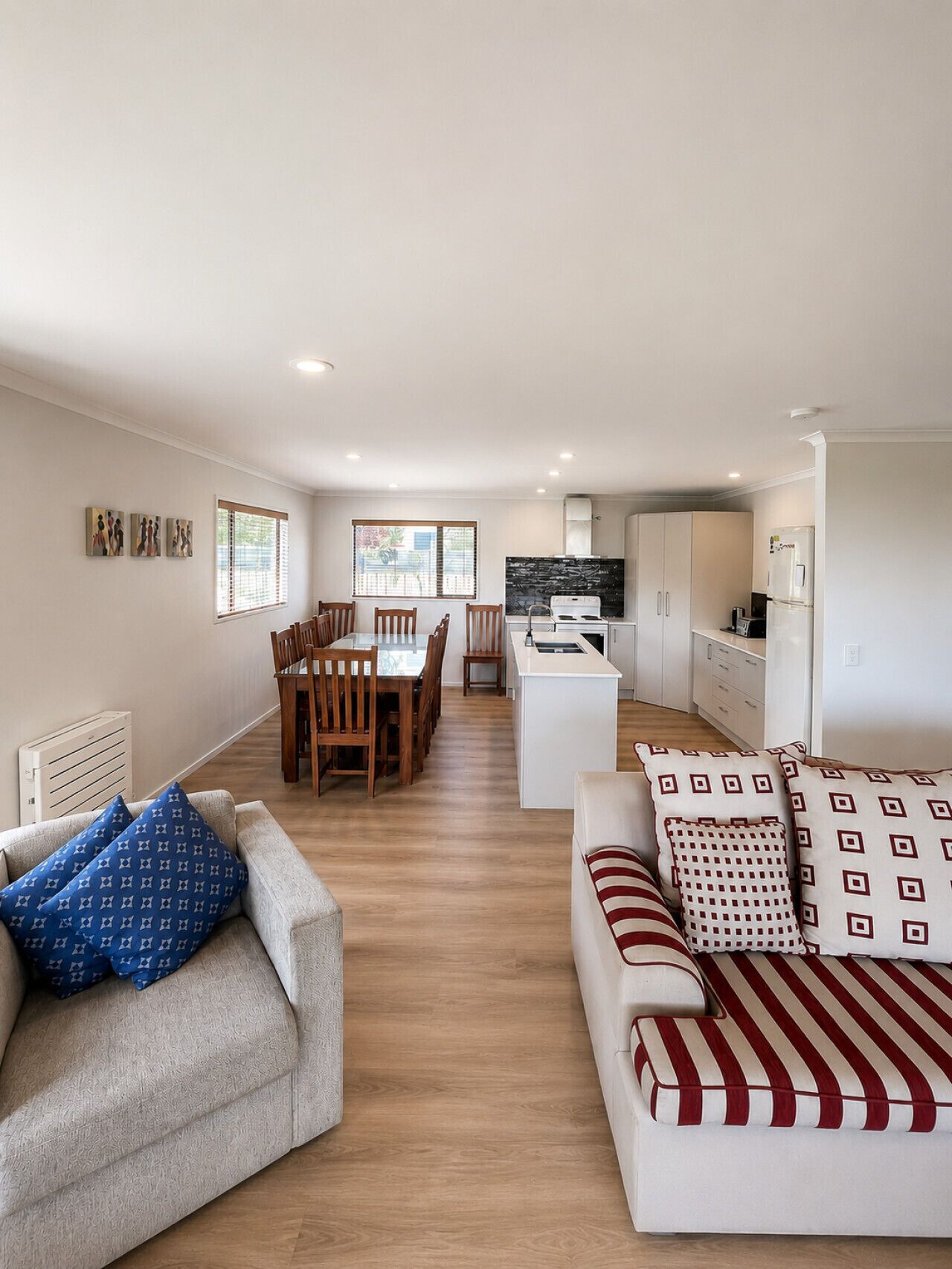 A modern living room with a sofa, a dining table with chairs, a kitchen area with cabinets and appliances, and a window with blinds.