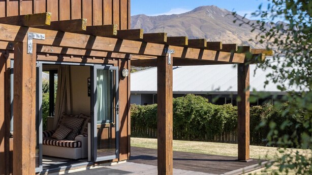 Une structure de pergola en bois, dotée d’une porte coulissante en verre qui donne accès à un espace détente agrémenté de coussins.