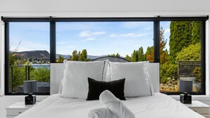Un dormitorio con una cama grande, ropa de cama blanca y vistas a un lago y árboles.
