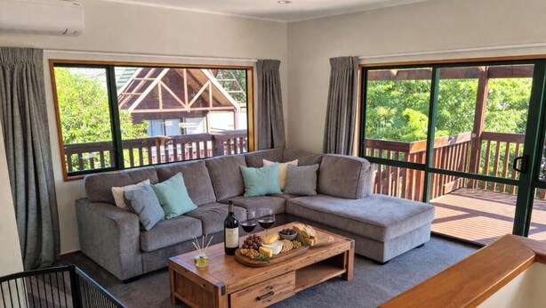 A living room with a sectional sofa, a wooden coffee table with food, and large windows with a deck view.