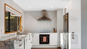 A modern kitchen with white cabinets, a stainless steel refrigerator, a built-in oven, and a window with blinds.