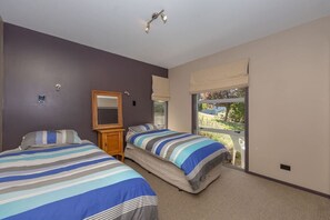 A bedroom with two beds, a wooden nightstand, a mirror, and a window with a view of greenery. - Wanaka Bach with Mountain Views (Wānaka)