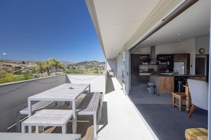 A balcony with a wooden table and chairs, a kitchen area with cabinets and a sink, and a view of a hilly landscape. - Wanaka Bach with Mountain Views (Wānaka)