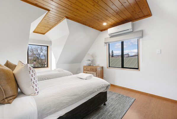 Un dormitorio con techo inclinado, una cama con ropa de cama blanca, una mesita de noche de madera y un ventanal con vistas a un campo.