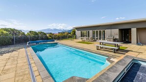 Une piscine rectangulaire bordée d’une terrasse carrelée, un espace patio avec une table et des chaises, et une vue sur les montagnes en arrière-plan.