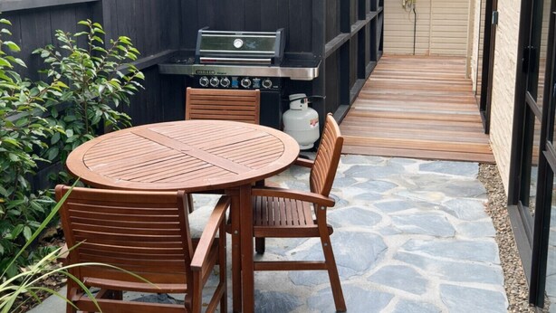 A patio with a round table and chairs, a barbecue grill, and a stone pathway leading to a house. - Wanaka Deluxe Studio (Wānaka)