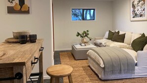 A modern bedroom with a bed, a wooden desk, a small stool, a window with a view of trees, and a painting on the wall. - Wanaka Deluxe Studio (Wānaka)