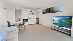 A modern living room with a flat-screen TV, a dining area with a small table and chairs, and a kitchen area with white cabinets and a built-in oven. - Studio Guest Home with Roys Peak Views (Wānaka)