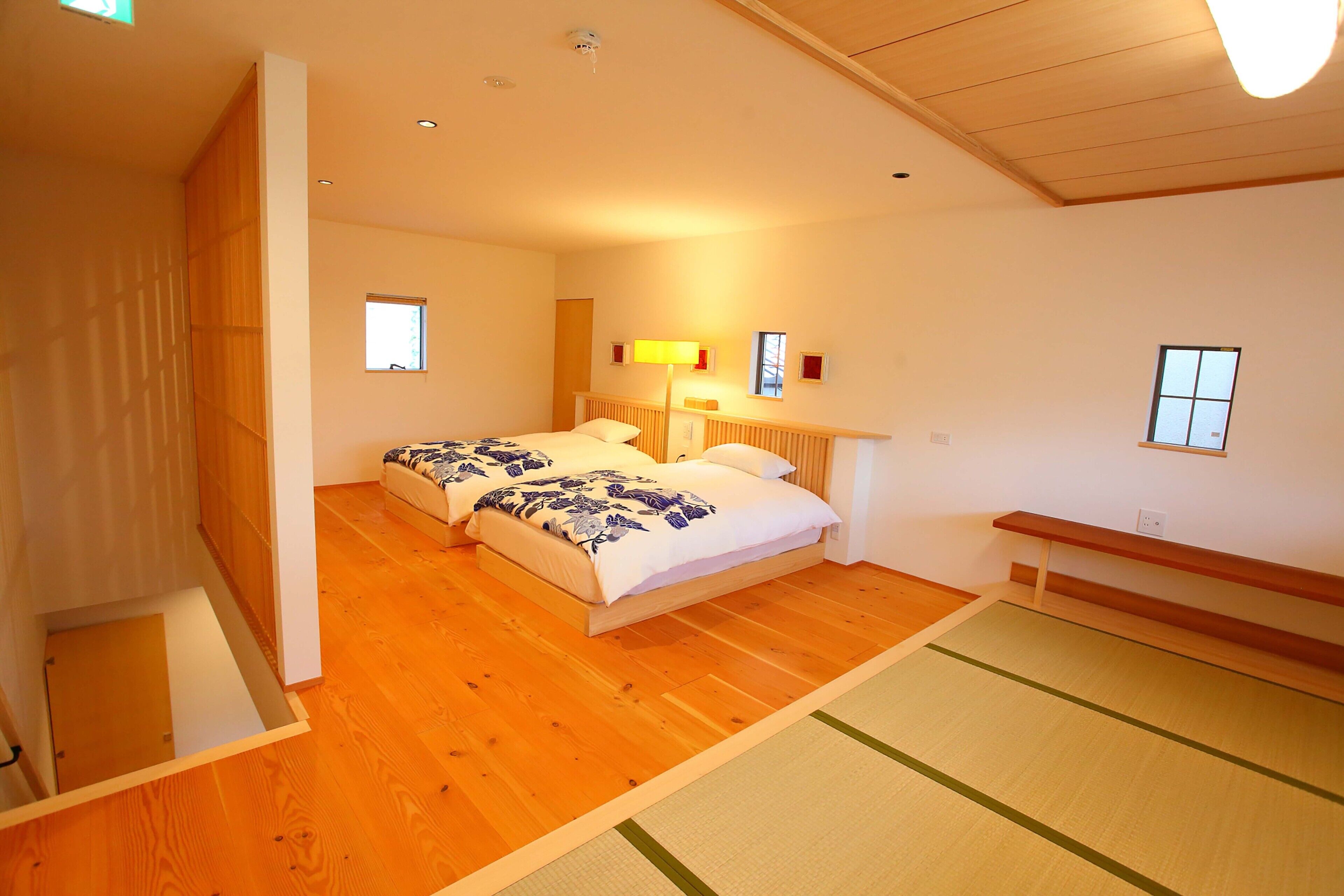 Quarto com duas camas, piso de madeira, esteira de tatame, ventilador de teto e uma janela pequena.
