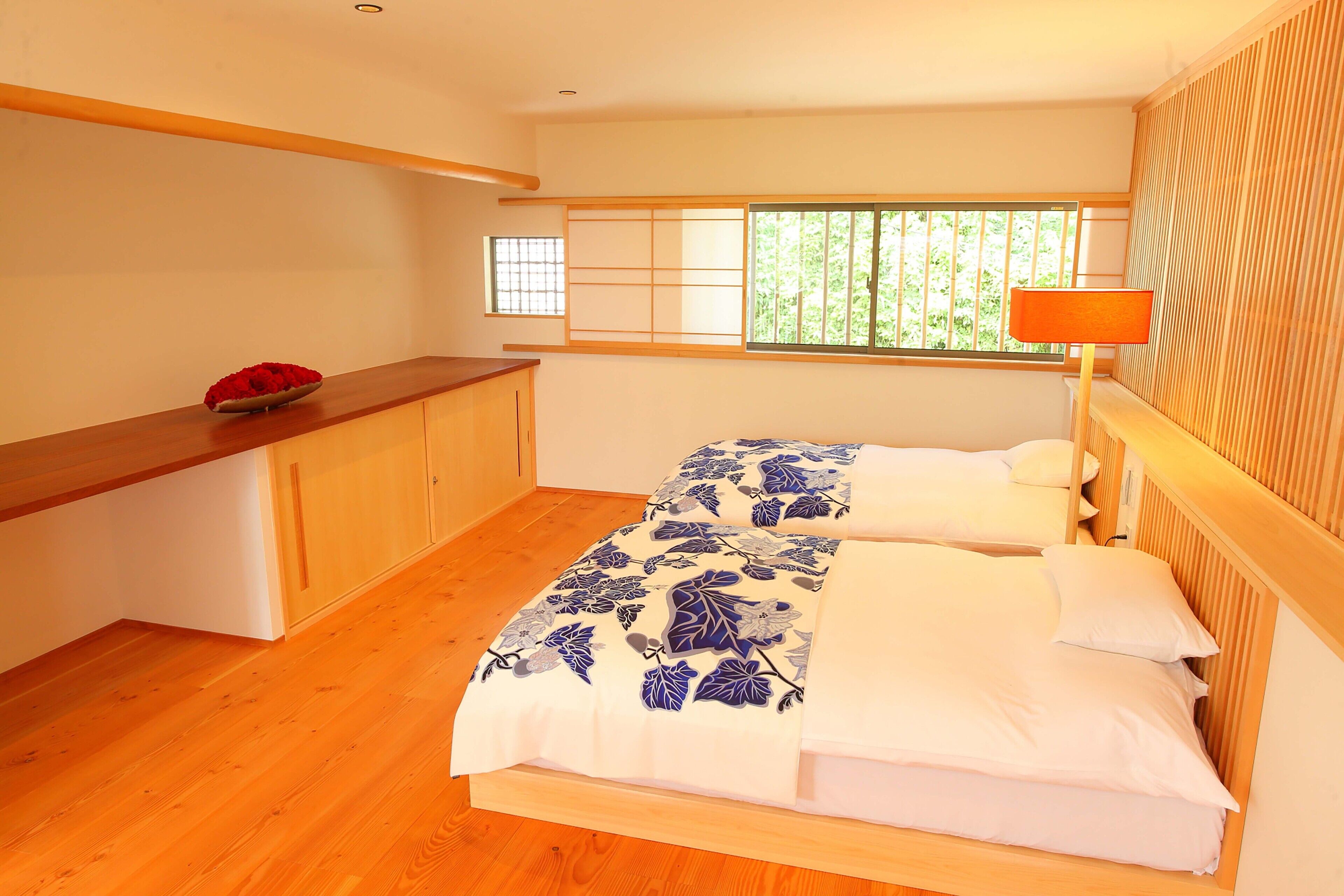 A bedroom with two beds, a wooden desk, a bowl of red flowers, and a window with a view of greenery.