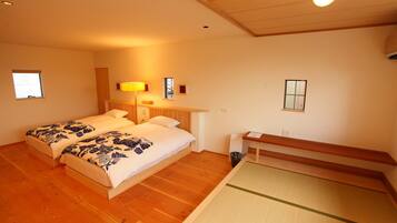 A room with two beds, a wooden floor, a bedside table, a lamp, and a small window.