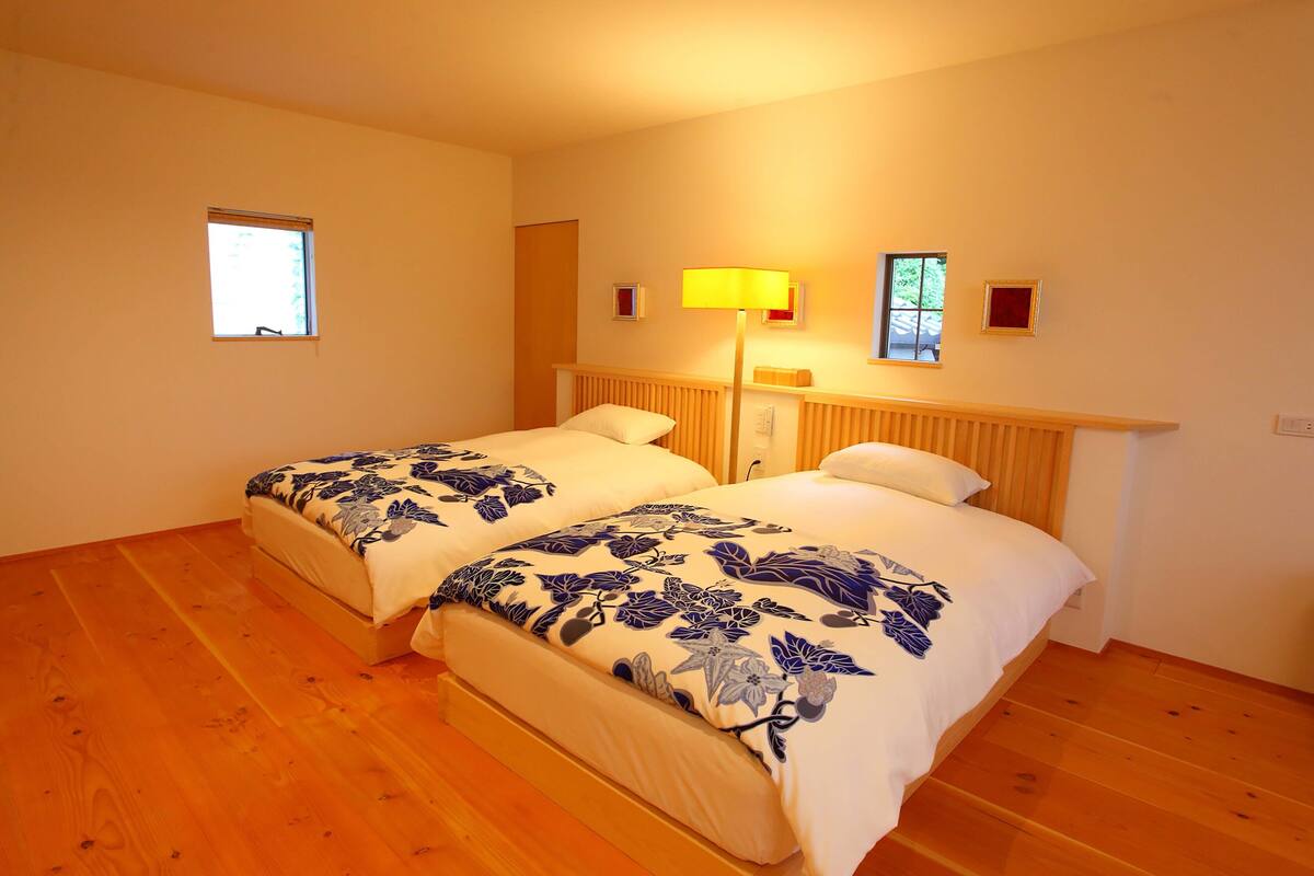 Une chambre avec deux lits, un plancher en bois, une fenêtre et une lampe fixée au mur.