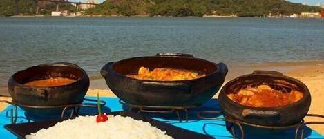 A table with three large pots of food and a plate of rice on a blue tablecloth by a body of water.