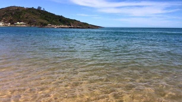 A beach with clear shallow waters, a distant hill with sparse vegetation, and a clear sky. - Hospedaria Oca Ruca (Guarapari)