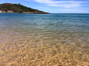 Une plage aux eaux claires et peu profondes, une colline lointaine à la végétation clairsemée et un ciel dégagé.