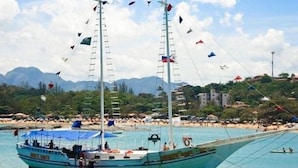 A boat with multiple masts and flags, docked near a beach with people and buildings in the background. - Hospedaria Oca Ruca (Guarapari)