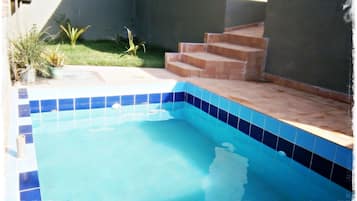 Una piscina exterior con azulejos azules, rodeada de un pequeño jardín y escalones que conducen a una zona cubierta.