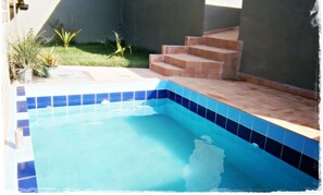 An outdoor swimming pool with blue tiles, surrounded by a small garden and steps leading up to a covered area. - Hospedaria Oca Ruca (Guarapari)