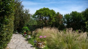 A stone pathway leading through a garden with various plants and a large tree in the background. - Escape to Serenita Villa in Arillas, Corfu - Private Pool, Sunset Views, and More (Arillas)