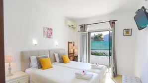 A hotel room with two beds, a bedside table, a lamp, a TV, and a window with curtains. - Renovated 2-Bed Gem in Kassiopi's Vibrant Tourist Village, WiFi, Terrace (Kassiopi)