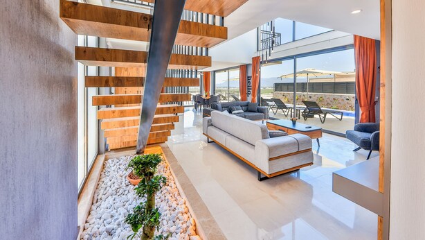 Un intérieur moderne avec un escalier en bois, un coin salon comprenant un canapé et des fauteuils, et une vue sur l’extérieur grâce à de grandes fenêtres.