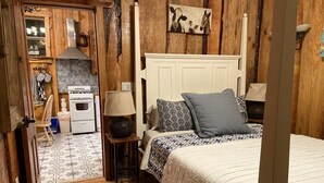 A cozy bedroom with a wooden floor, a white bed with patterned pillows, a bedside table with a lamp, and a view into the kitchen with a stove and tiled backsplash. - Pacific Coast Highway Milkhouse (San Luis Obispo)