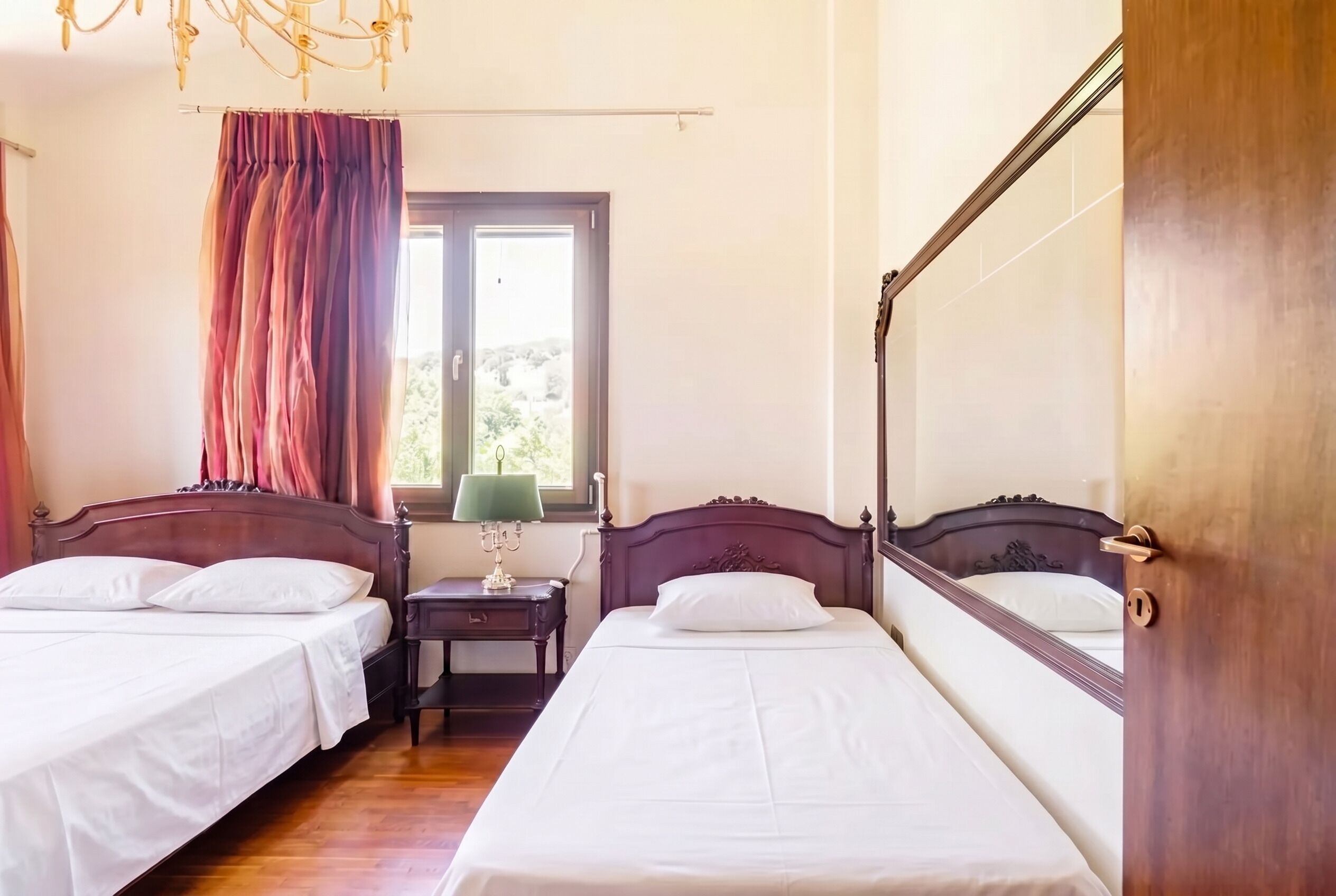 A hotel room with two beds, a window with curtains, a nightstand with a lamp, and a wooden door.