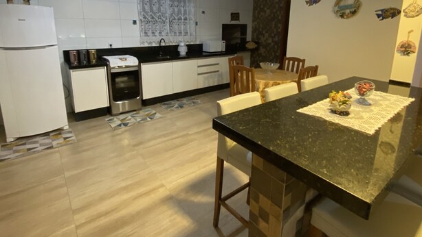 A modern kitchen with white cabinets, a black countertop, and a dining area with a table and chairs. - Casa de Praia e Campo com Piscina (Garopaba)