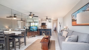 A modern living room with a grey sofa, wooden dining table, and kitchen in the background. - Great Location! Walk to Everything in South Kihei! (Kihei)