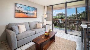 A living room with a grey sofa, a wooden coffee table with fruit, a painting of a beach, and a balcony with a table and chairs. - Great Location! Walk to Everything in South Kihei! (Kihei)